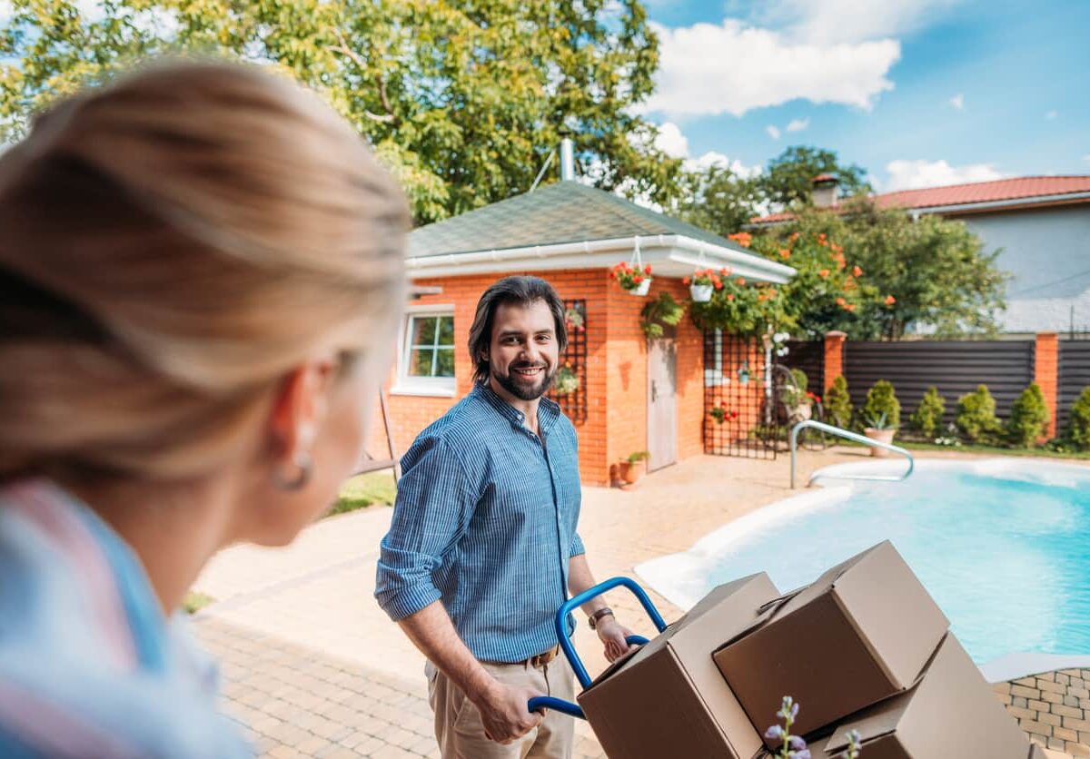 Man moving stacked boxes on a moving cart, with a pool in the background and a woman’s head in the foreground.