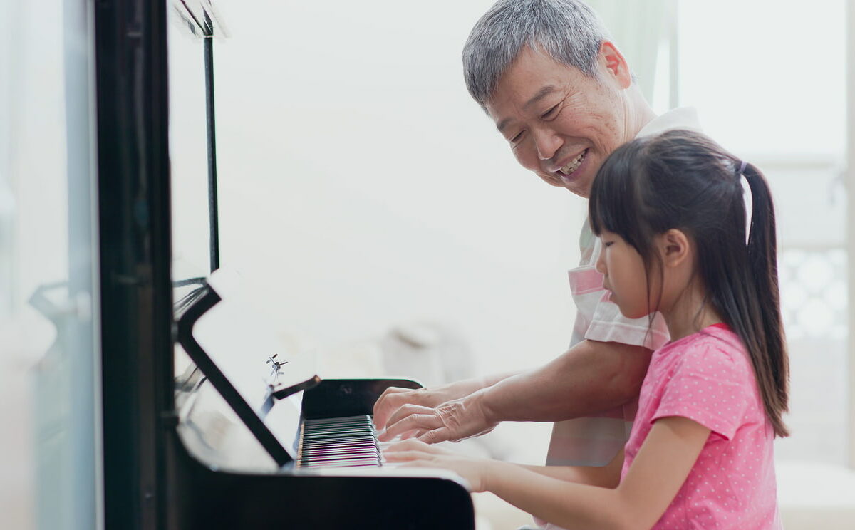 Elderly man smiles while playing piano with young girl.