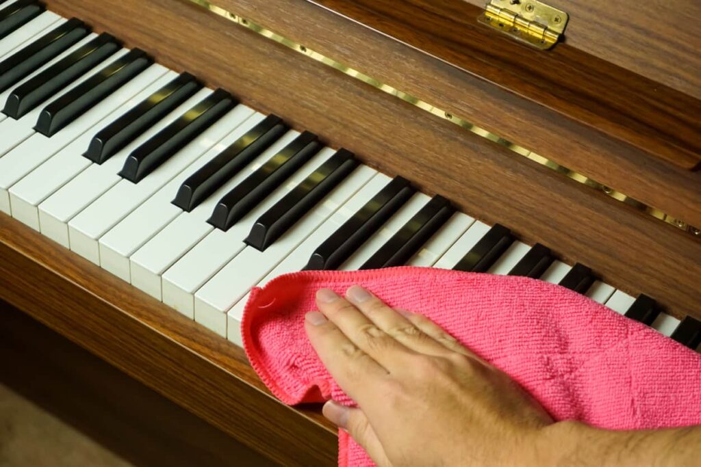 A hand wipes down the keys of a piano using a pink microfiber cloth.
