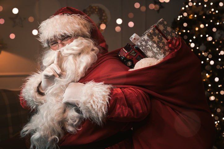 Smiling Santa with index finger to his lips, making a shushing gesture.