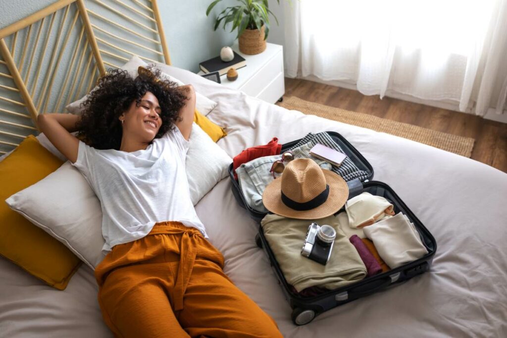 A happy woman lying on the bed next to an open suitcase full of clothes, a camera, a passport, and a hat, ready to go on a summer vacation.
