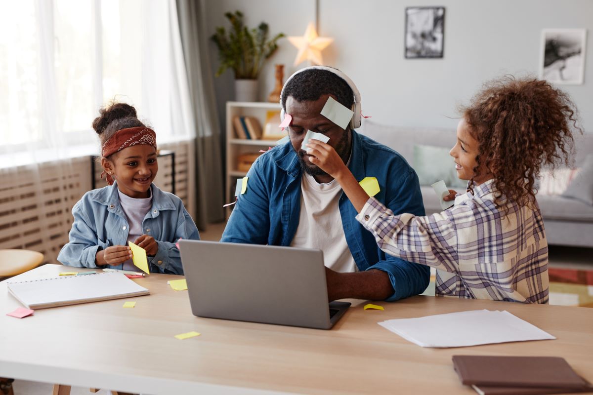 A portrait of two happy children sticking notes on their father trying to work from home.